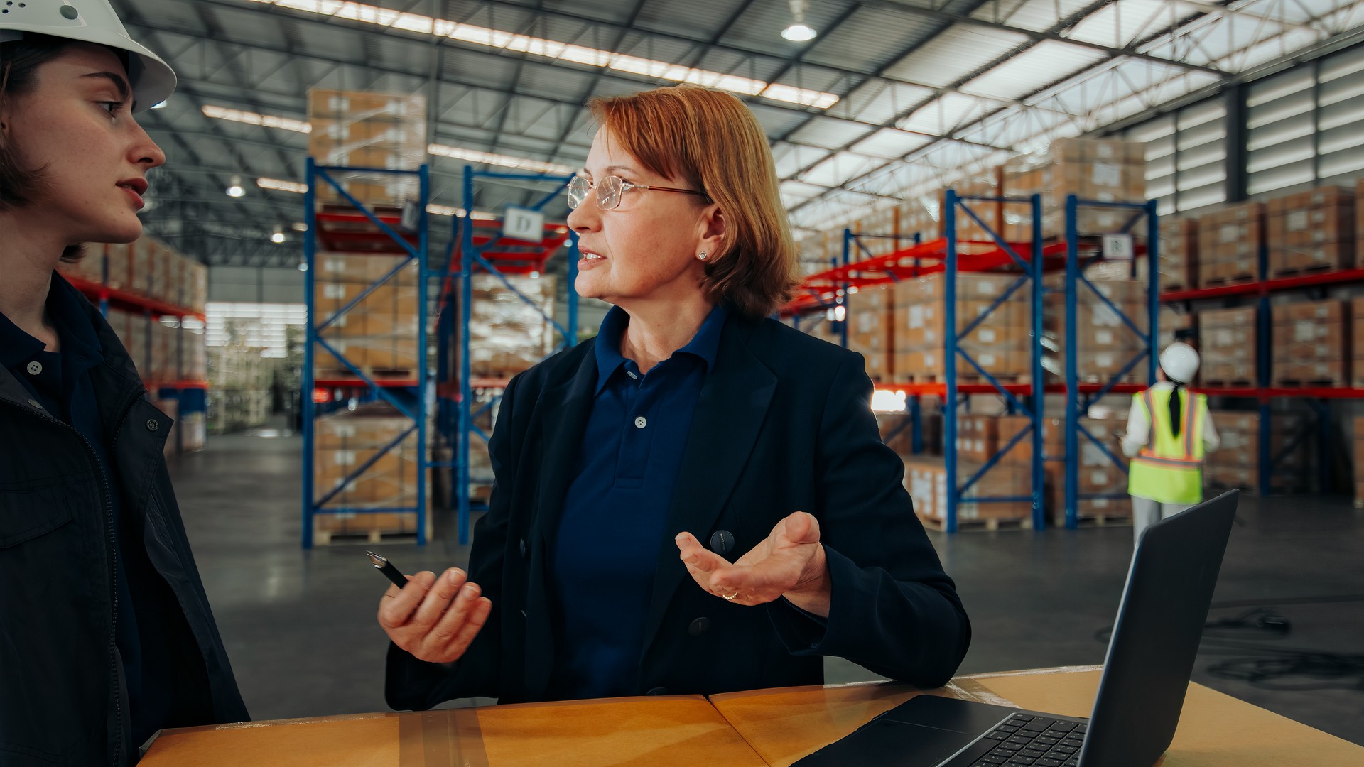 A senior logistics manager and a warehouse operative are deeply engaged in a workflow optimization discussion using a laptop in the warehouse